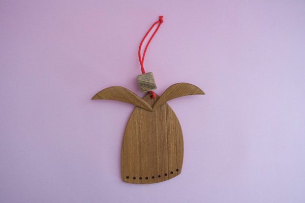 Angel iz tikovega lesa je okras v obliki angela z ukrivljenimi krili, majhnimi luknjicami ob robu, rdečo vrvico ter lesenim perlom. Postavljen je na svetlo rožnato ozadje.