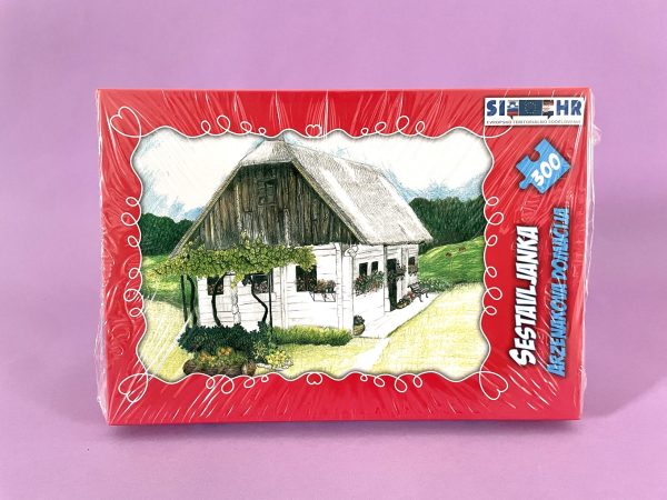 Sestavljanka iz 300 delov z naslovom "Sestavljanka - Arzenakova domačija" ima ilustracijo kmečke hiše v zeleni pokrajini. Rdeča škatla ima belo oblogo, besedilo in nalepko z napisom "Sestavi Junka Kmeckim Domaca" in "SI-HR.