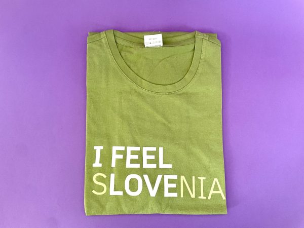 Majica I FEEL SLOVENIA, olivno zelena majica z belim napisom "I FEEL SLOVENIA" na sprednji strani, je prikazana zložena na vijoličnem ozadju.