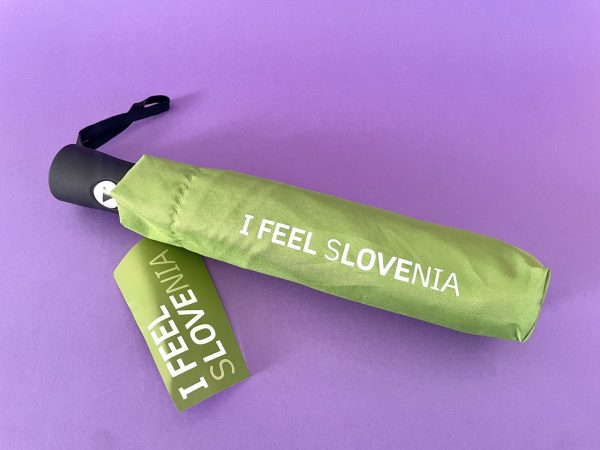 Zeleni dežnik I FEEL SLOVENIA s sloganom, natisnjenim na naslovnici, stoji na vijolični površini, spremlja pa ga ustrezna zelena etiketa z enakim besedilom.