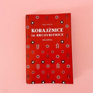 Knjiga "Korajžnice in brkovritnice" avtorja Toma Costa z rdečim ovitkom, okrašenim z belimi, črnimi in modrimi cvetličnimi in geometrijskimi vzorci, je razstavljena na svetlo rožnati podlagi.
