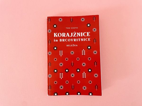 Knjiga "Korajžnice in brkovritnice" avtorja Toma Costa z rdečim ovitkom, okrašenim z belimi, črnimi in modrimi cvetličnimi in geometrijskimi vzorci, je razstavljena na svetlo rožnati podlagi.