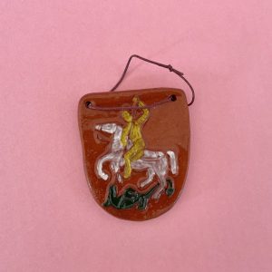 Magnet Šentjurski grb je rjav keramični magnet v obliki ščita z rumeno figuro na belem konju s kopjem nad zelenim zmajem na rožnati podlagi. Skozi dve zgornji luknjici je navlečena temna vrvica.