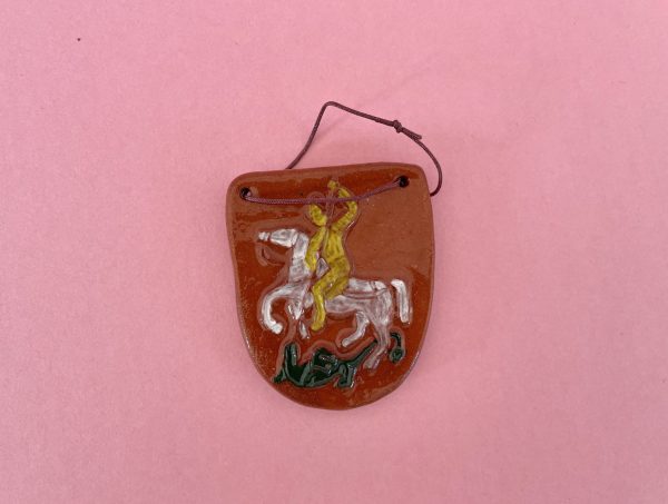 Magnet Šentjurski grb je rjav keramični magnet v obliki ščita z rumeno figuro na belem konju s kopjem nad zelenim zmajem na rožnati podlagi. Skozi dve zgornji luknjici je navlečena temna vrvica.