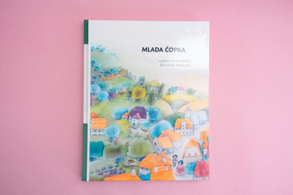 Knjiga "Mlada čopka" leži na rožnati površini, na naslovnici pa je živahna ilustracija vasi s hišami, drevesi in majhnimi figurami.
