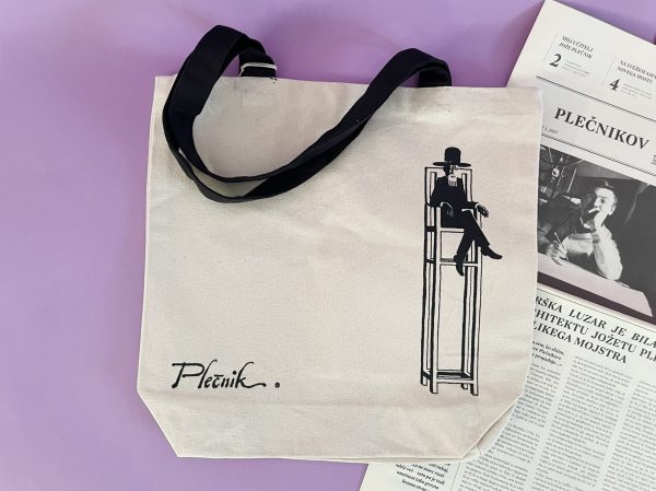 Vrečka Plečnik je bež torba s črnim ročajem z minimalistično ilustracijo sedeče osebe na visokem stolu in napisom "Plečnik" pod njo. Prikazana je poleg časopisa na sivkasti površini.