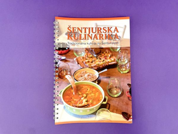 Na naslovnici kuharske knjige Šentjurska kulinarika so na vijoličnem ozadju lonec juhe, skleda riža in pekač na mizi s kozarci.