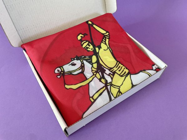 Šentjurska zastava, prepognjena rdeča tkanina z rumenim bojevnikom na belem konju, je razstavljena v odprti beli škatli na vijolični površini.