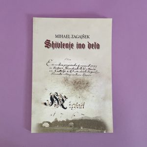 Na rožnati površini je razstavljena knjiga z naslovom "Mihael Zagajšek, Shivlenje ino delo". Na njenem ovitku so vidni staro ročno napisano besedilo, okrasna inicialka in šibke podobe hiš na dnu.