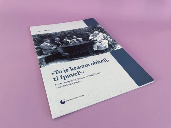 Knjiga z naslovom "To je krasna obitelj, ti Ipavci!" ima na naslovnici črno-belo fotografijo ljudi za mizo in je razstavljena na rožnati površini.