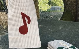 Kuhinjska krpa z noto, bela brisača s teksturo vaflja z rdečimi ali zelenimi notami, visi in sedi zložena na kamniti površini na prostem, zvezana z vrvico v bližini drevesa.